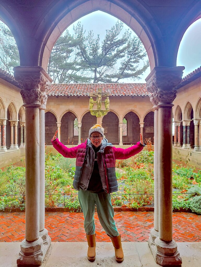 View of me in my winter coating with my hands up and I a standing in between two columns surrounding an outdoor garden at the MET clositers. 