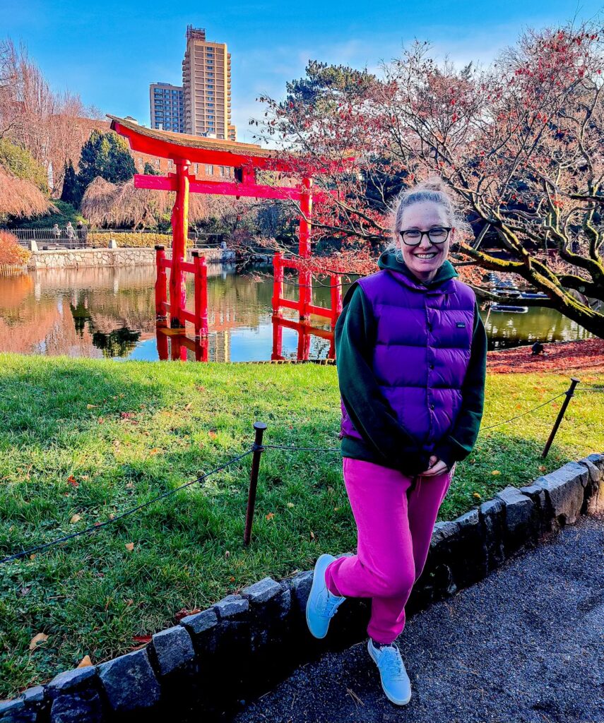 Me standing in a purple vest and green sweatshirt with pink pants in front of a red Japanese gate at the Brooklyn Botanic Garden. 