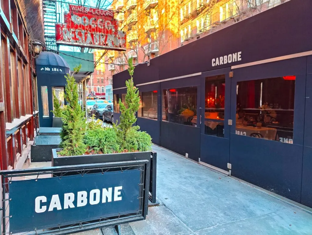 The exterior of Carbone. You can see a red sign above green potted plants and blue signs on the ground. There is also a square tent with tables and "carbone" on it for outdoor dining. 