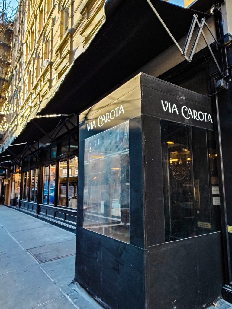 There is a black awning above a black, plastic box with "Via Carota" in white letters. It sits outside of the entrance to the restaurant to protect people indoors from the cold in the winter. 