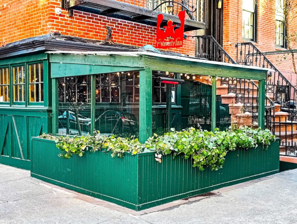 The exterior of the Waverly Inn sits on the ground floor of a historic, brick building. It is an enclosed green porch with plants that sit in window boxes. around the exterior of the restaurant. A red, "Waverly Inn" sign sits at the top. 