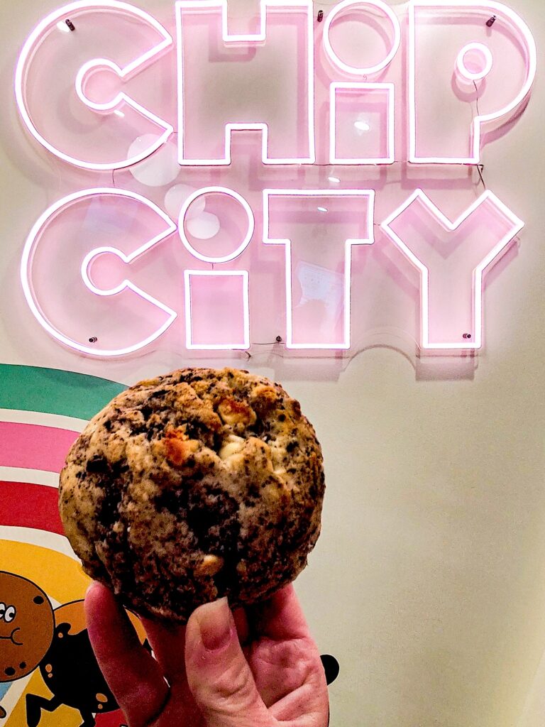 A view of me holding a chip city chocolate chip cookie in my hand. It am holding it in front of a neon chip city sign with pink letters. This is one of the best chocolate chip cookies in NYC. 