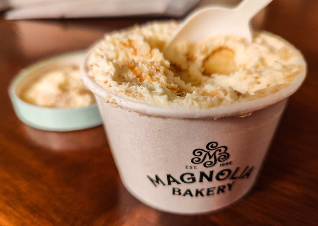 A view of a small container of banana pudding from Magnolia bakery on a wood table. I have my plastic spoon in the container and  the top on the table. 