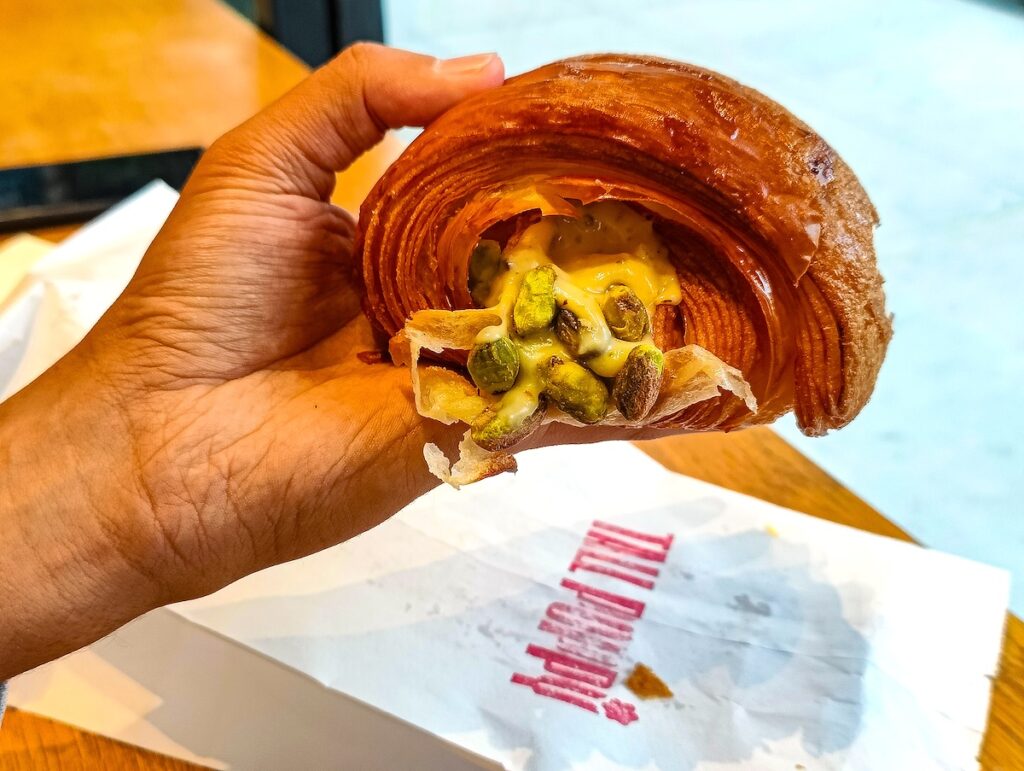 My partner holding a croissant. You can see the middle bursting with pistachios and a whit bag with red letters that say "Tall Poppy" sitting on a wood table. 