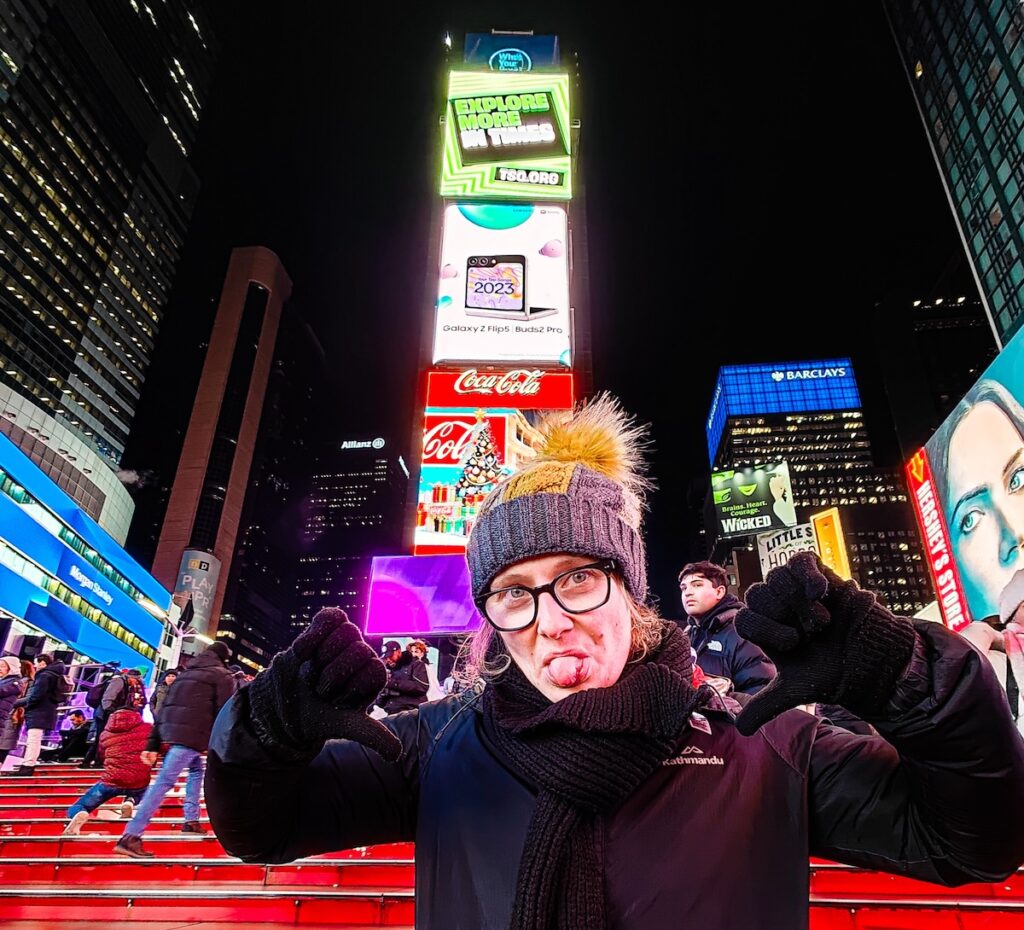 A picture of my sticking my tongue out with my hands up by my face at night in Times Square after staying at one of the best hostels in NYC. 