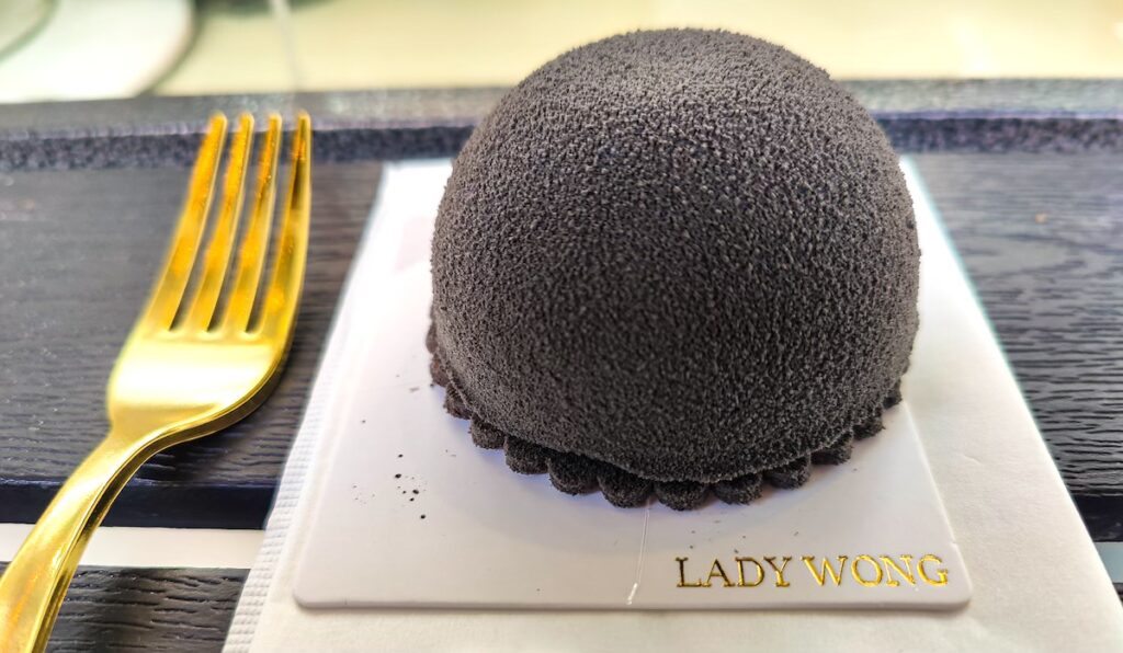 A black dome that is a cake from lady Wong's. You see a gold fork on the left and the cake sits on a white board on a black table. 