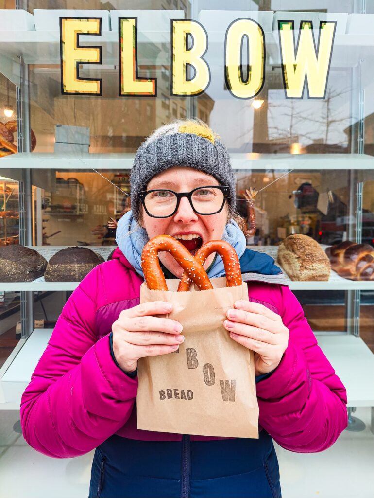 Me wearing a gray winter hat and a purple winter jacket while holding a pretzel in a brown bag and standing in front of Elbow Bakery. 