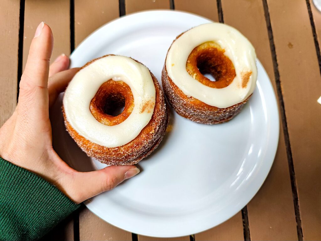 Two cronuts on a white plate with whtie frosting on top. I am holding one cronut with my hand while wearing a green sweatshirt.  