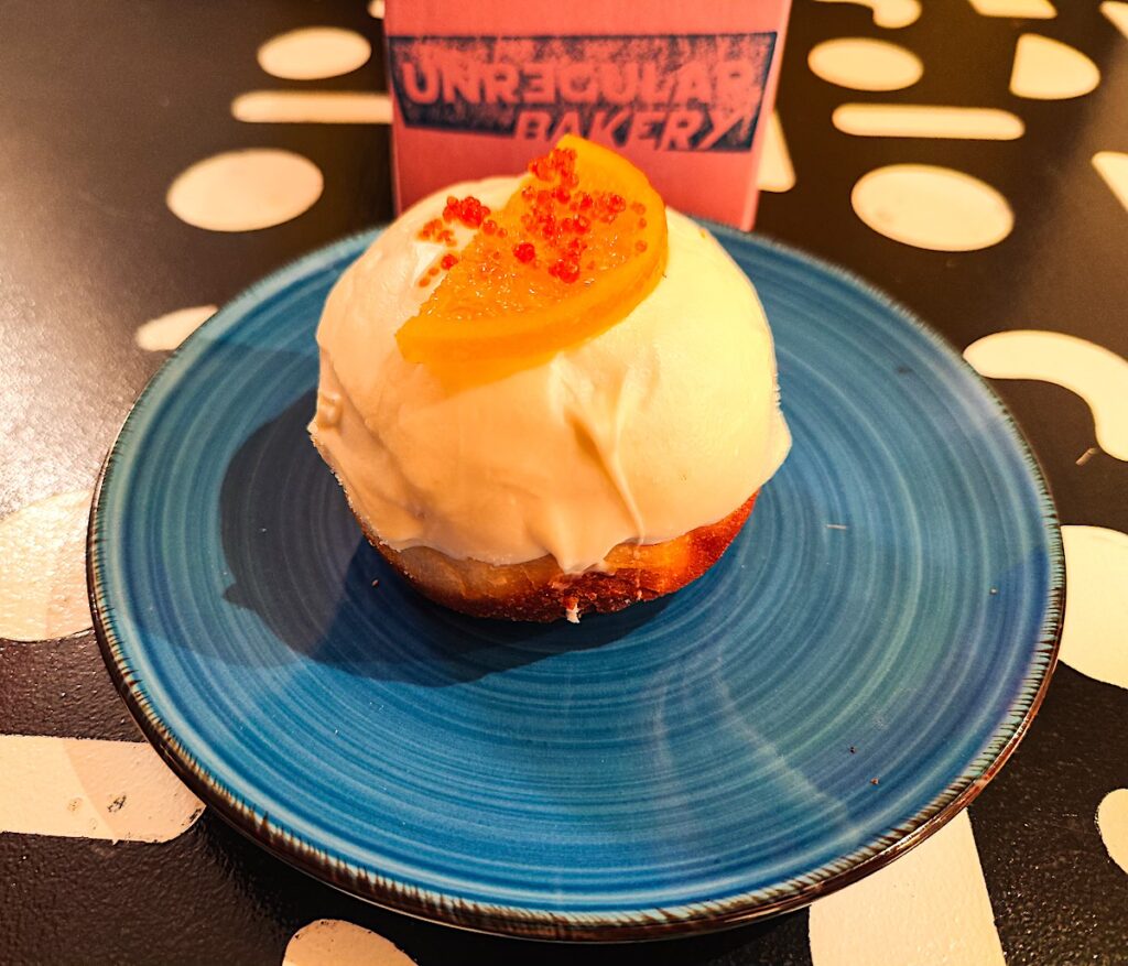 A view of a round desert with white frosting on top and a candied orange slice with lots of orange pearls. The dessert sits on a blue plate with a pink box in back and on a black table. 