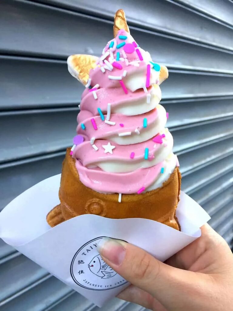 View of me holding a fish bread with pink and white ice cream and ears/unicorn horn in front of a gray gate at a store. 