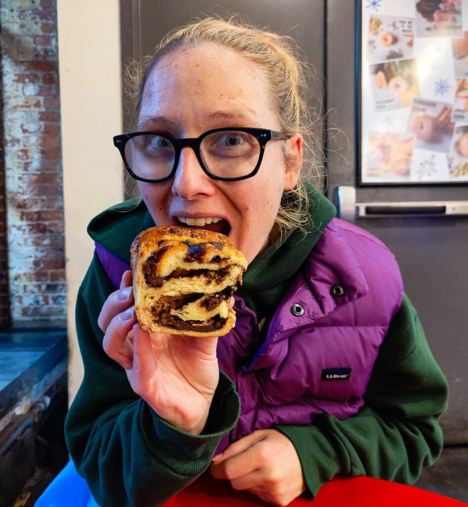 Me in black glasses and holding a giant piece of chocolate babka as I bite into it in a green sweatshirt and purple vest. 