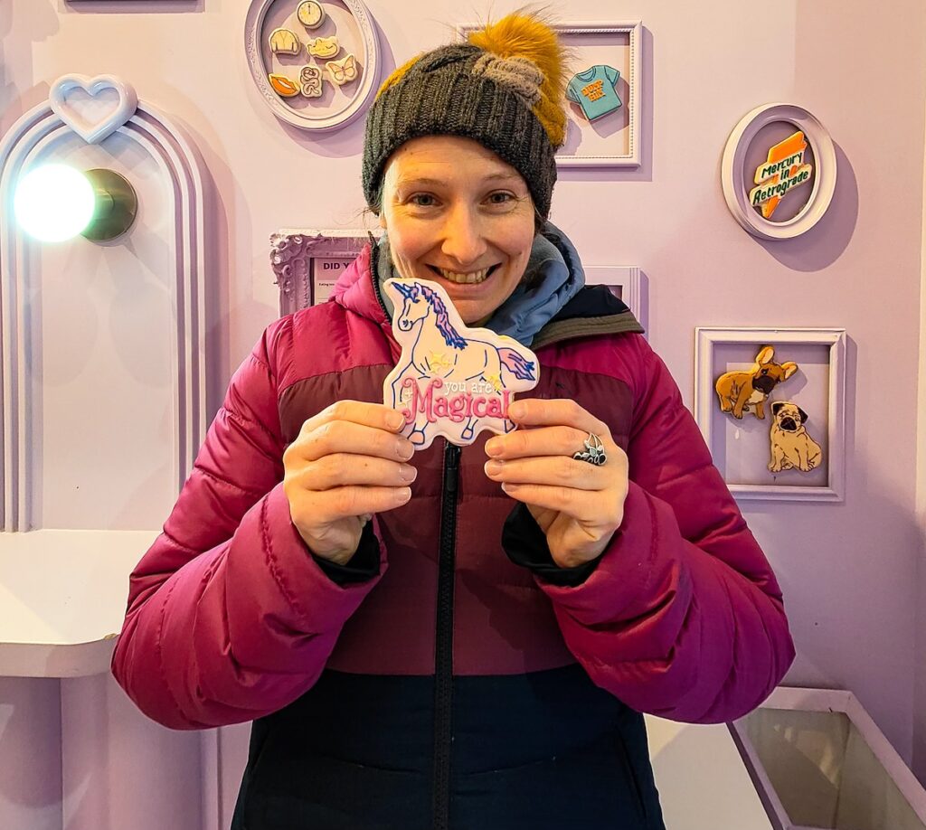 View of me pin a pink/purple winter coat and gray hate with a yellow pom pom. I am holding a Funny Face cookie that says "magical" with a unicorn on top. 