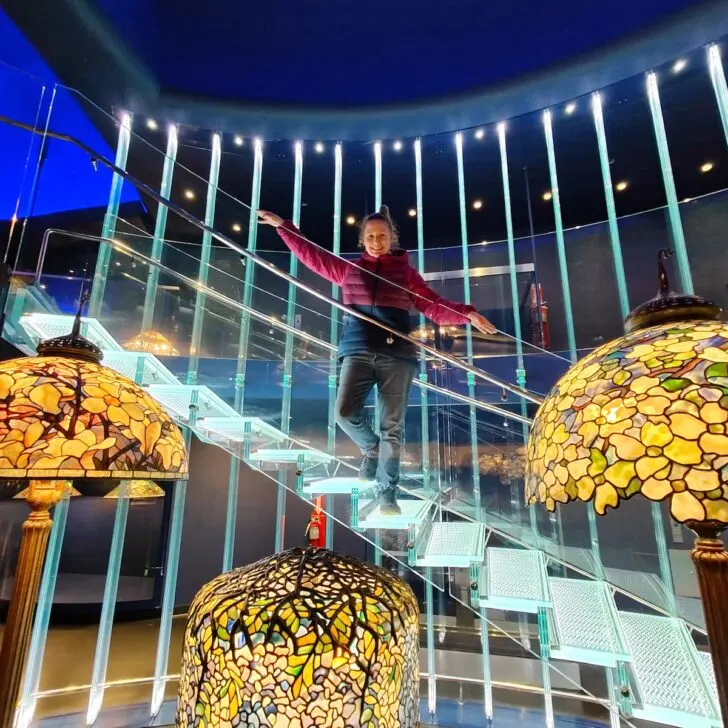 View of me standing ona. glass stairway in front of three tiffany lamps on display in the New York Historical Society.