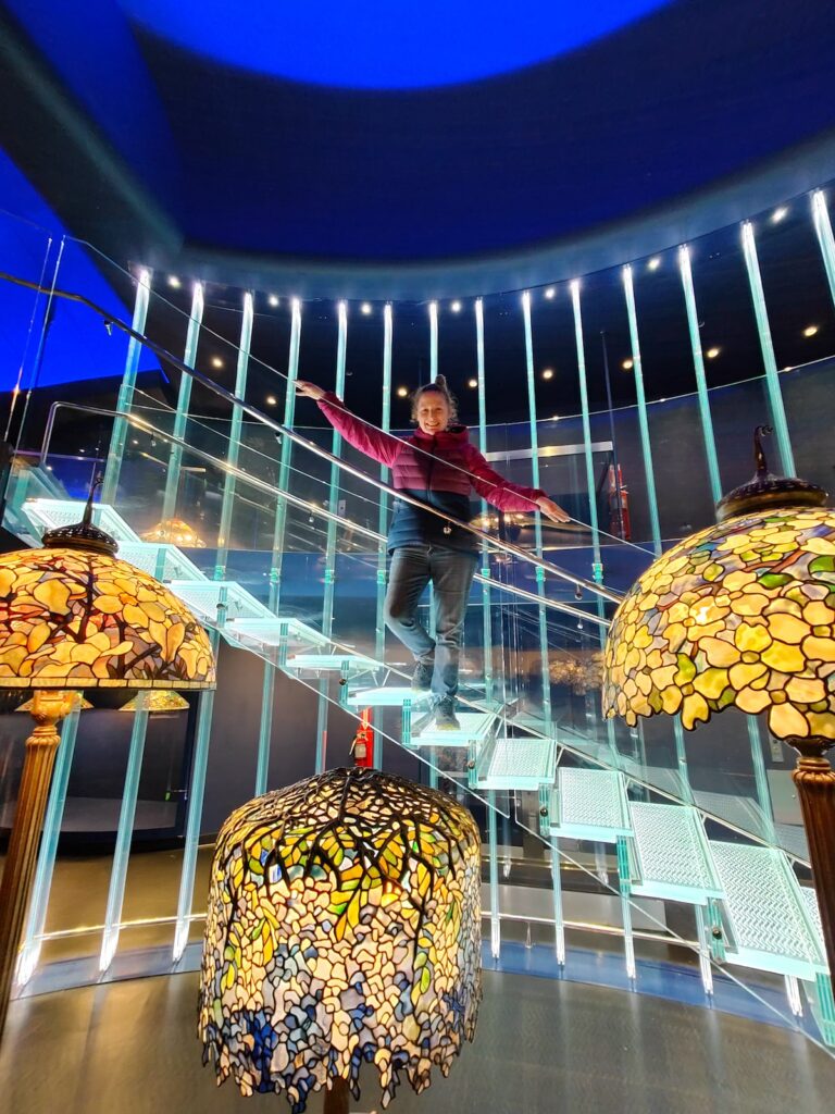 View of me standing ona. glass stairway in front of three tiffany lamps on display in the New York Historical Society. 