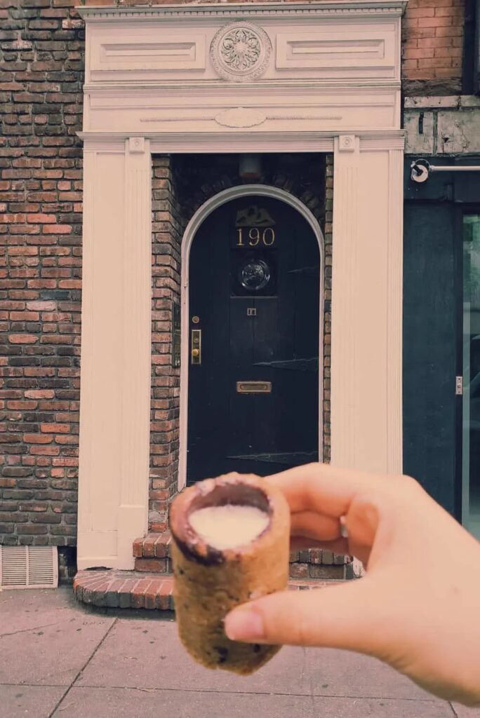 A view of me holding a cookie and milk shot from Dominique Ansel Bakery. My hand is in front of a white door and you can see a chocolate chip cookie shot glass filled with vanilla-infused milk. 