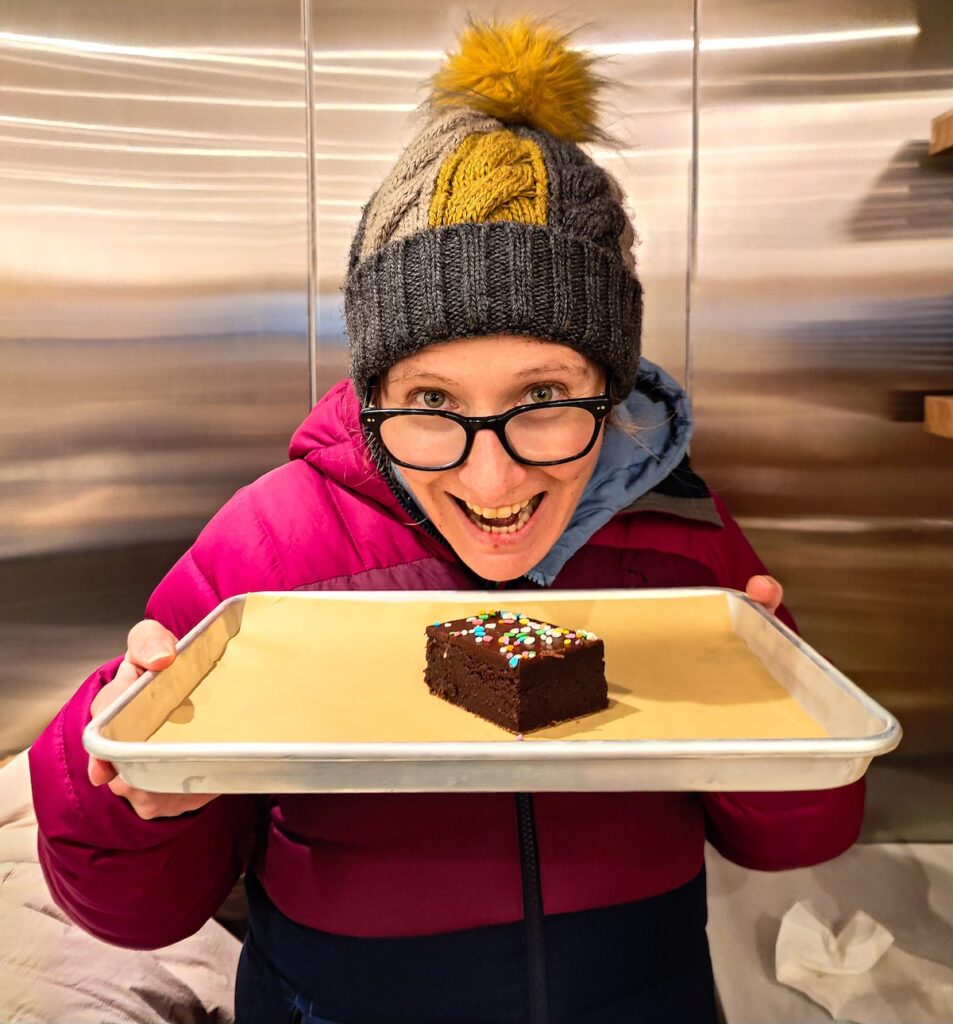 Me wearing glasses, a purple coat and a winter hat as I hold a metal tray with a brownie on it from Hnai's in NYC. 