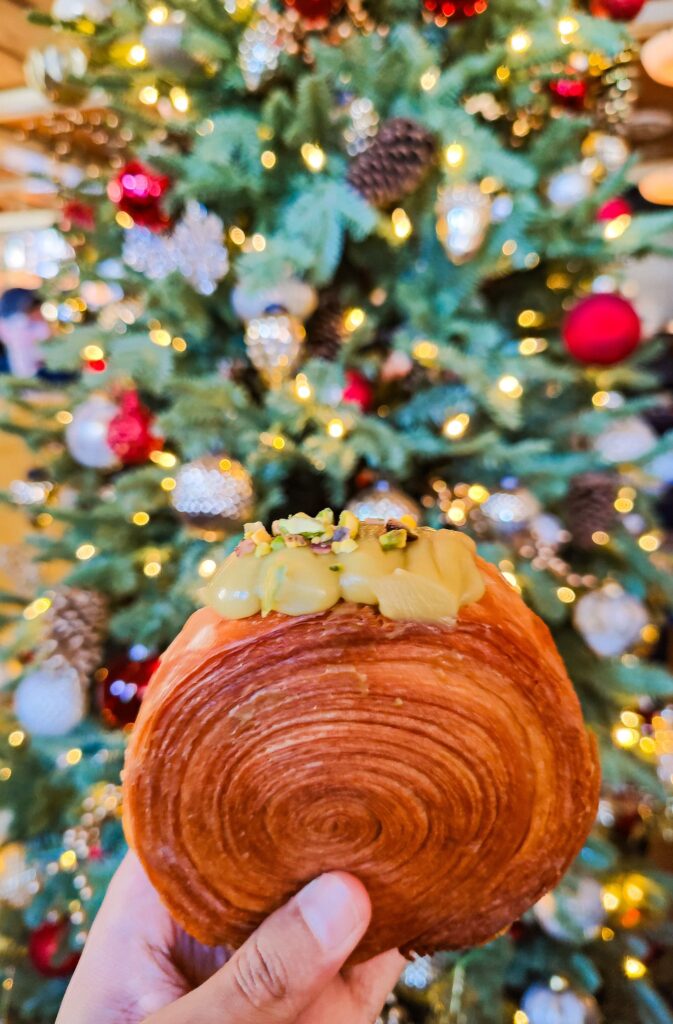 Holding a round croissant with green icing on top in front of a Christmas tree. 