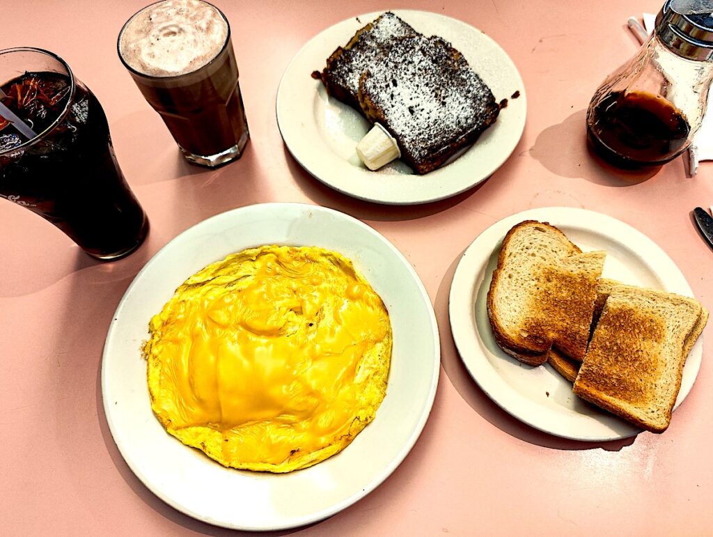 Aerial view of an omelet, toast, and banana nut french toast on a table with an egg cream. 