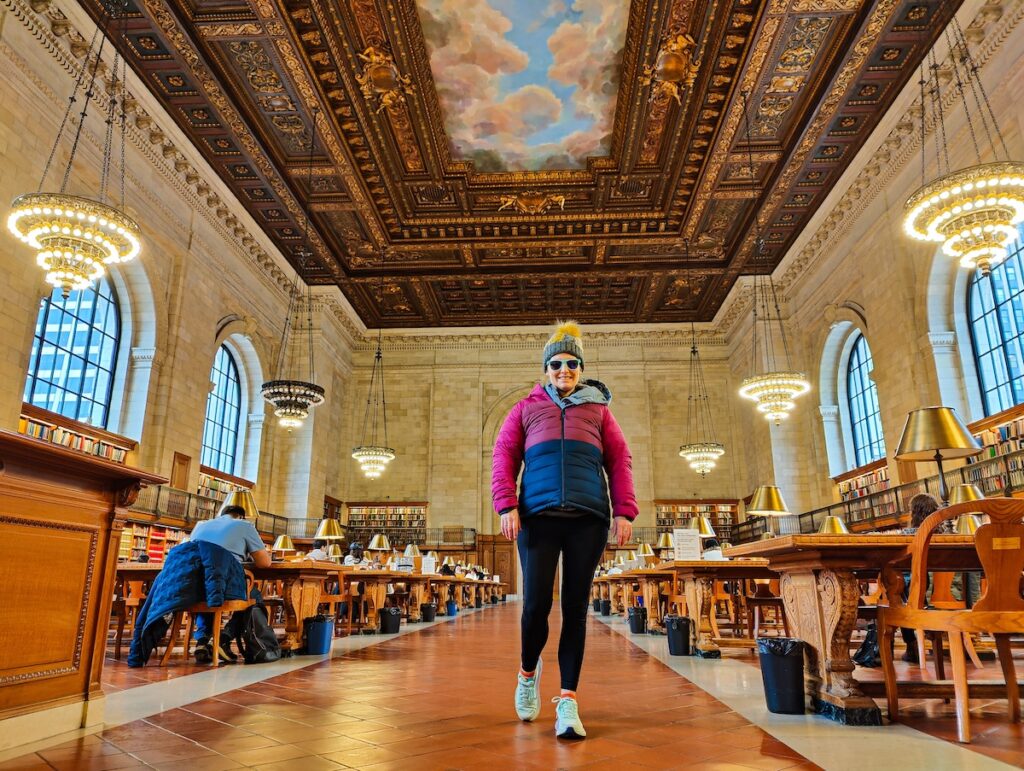 A view of me in a winter jacket and a gray hat walking in tights and turquoise sneakers down a walkway in the reading room in the New York Public Library. 