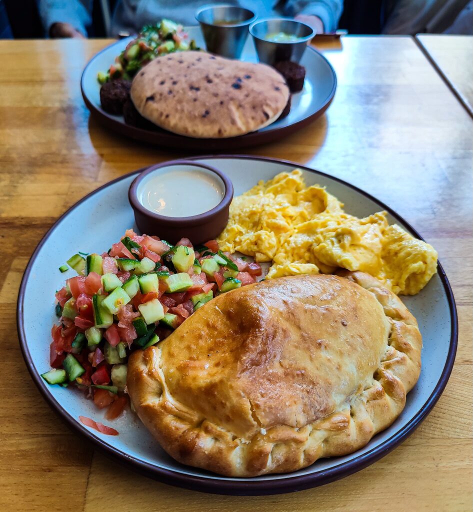 Aerial view of a plate fileld with bread, scrambled eggs, salad, and sauce. It sits on a wooden table and their is another plate in the brackground.  