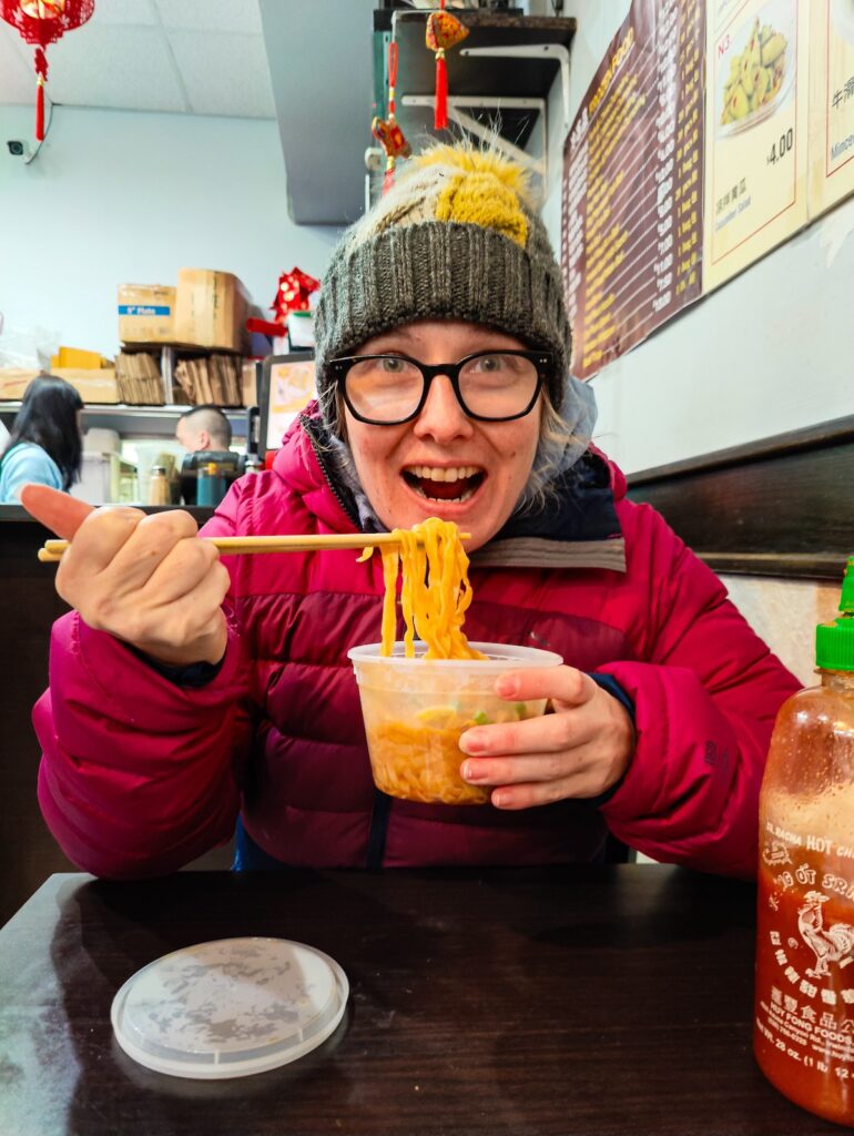 View of me eating peanut butter noodles from a restaurant in Chinatown with a winter hat and jacket on.  I am eating in NYC in March. 