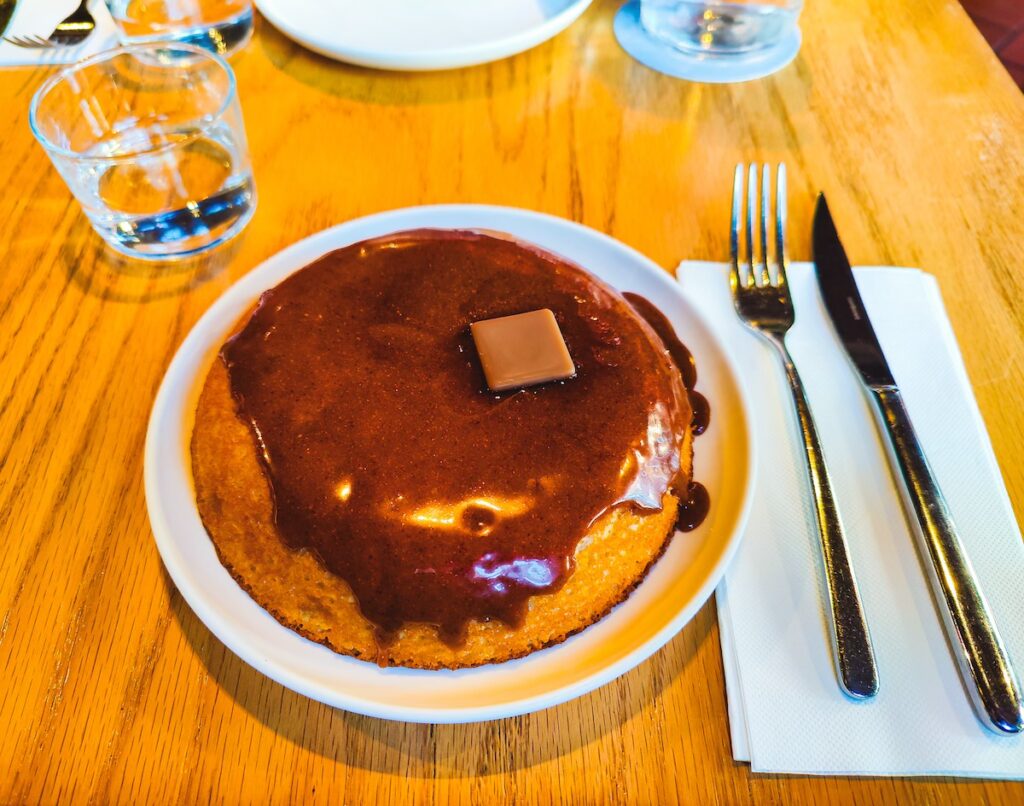 Aerial view of a thick pancake on a white plate with chocolate sauce on top with a pat of butter. You can see a napkin on the right hand size with a knife and fork. 