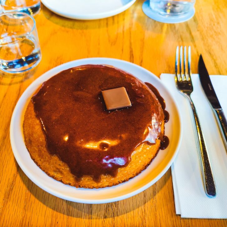 Aerial view of a thick pancake on a white plate with chocolate sauce on top with a pat of butter. You can see a napkin on the right hand size with a knife and fork.