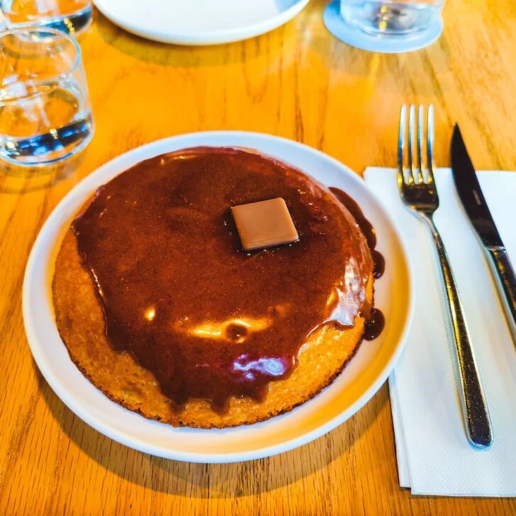 Aerial view of a thick pancake on a white plate with chocolate sauce on top with a pat of butter. You can see a napkin on the right hand size with a knife and fork.