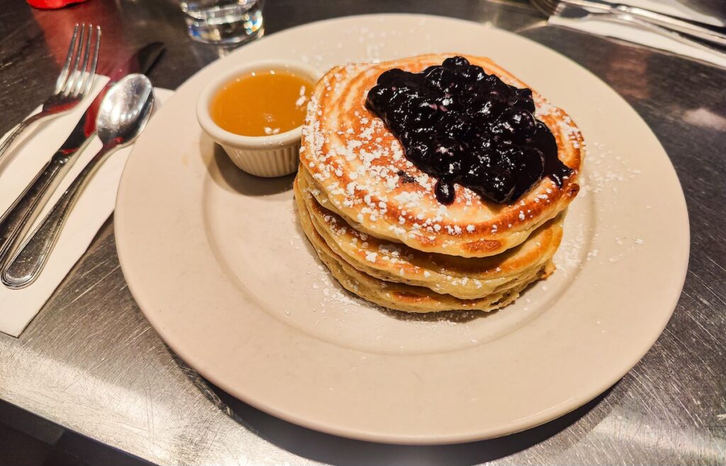 Aerial view of a plate filled with pancakes and maple butter on the side. The pancakes are topped with blueberry sauce and powdered sugar and sit on a white plate. 