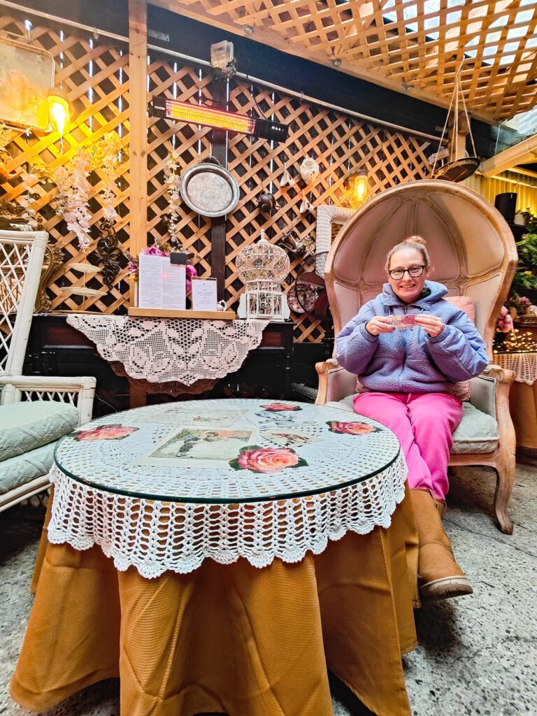 View of my sitting at a table inside of Brooklyn High and Low. I am in pink pants and a blue fleece and I am holding a cup of tea as aI sit around a small round table. 