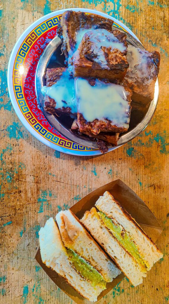 Aerial view of chocolate french toast smothered in white glaze on a Malaysian-style plate with kaya toast on another plate on a distressed wood table. This is a pick for the best french toast in NYC. 