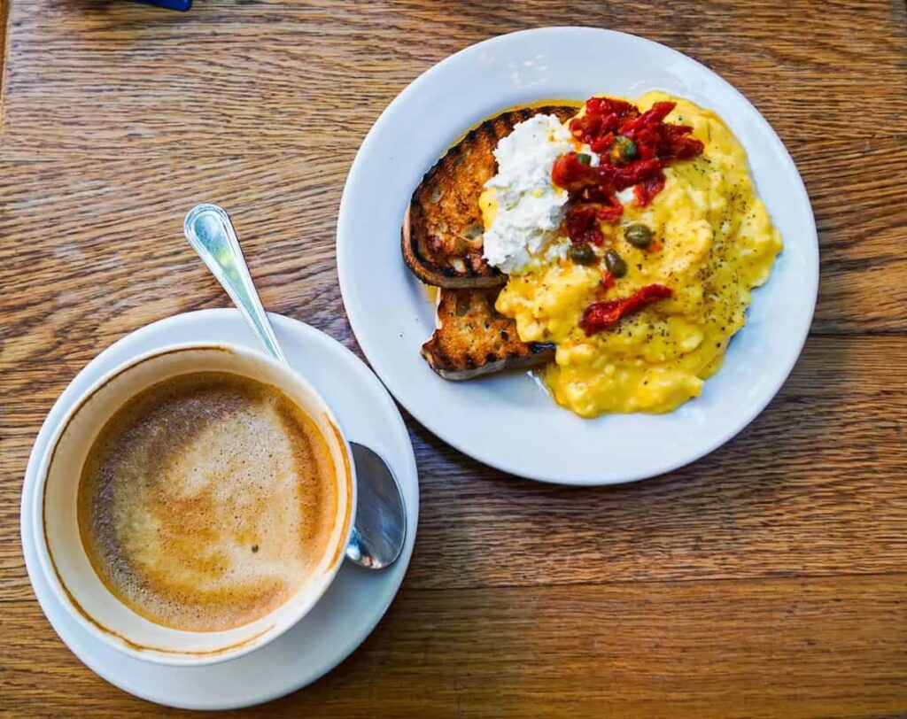 Aerial view of a half cup of coffee with milk and a small plate with toasted bread, scrambled eggs, and cheese/red peppers on top from Buvette. 