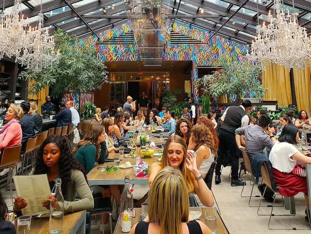 A view of the dining room in Nomo Kitchen. You can see large chandeliers hanging from the glass ceiling as well as graffiti hearts in the background. You see people sitting at the tables as they enjoy the best pancakes in NYC. 