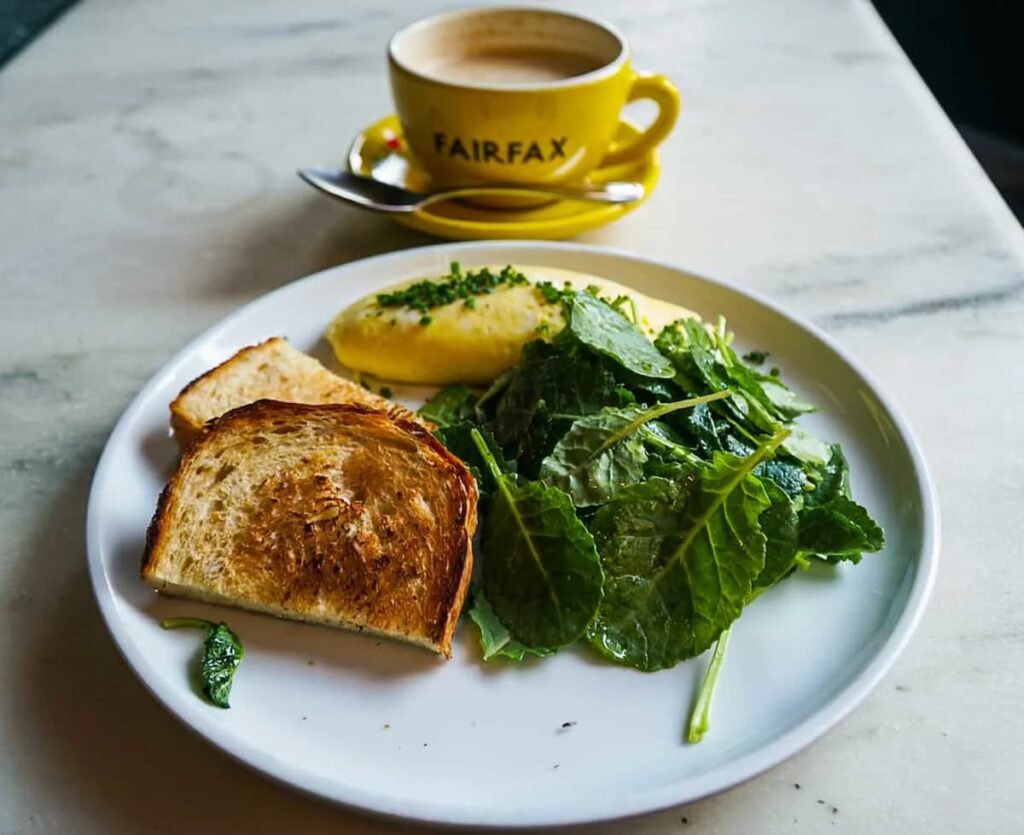 View of breakfast at Fairfax. You can see a yellow mug of coffee in the back with a plate full of eggs, kale salad, and toasted bread in the front. 