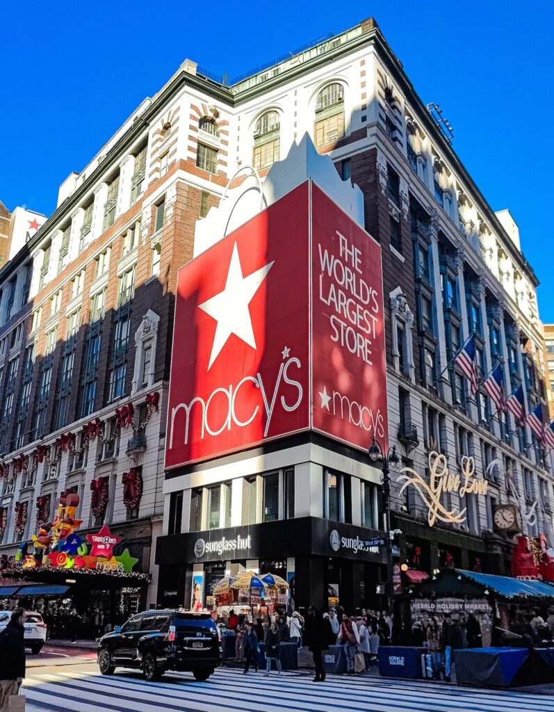 View of Macy's in Herald Square on a sunny day. You can see people walking by and a huge red shopping bas that reads "Macy's, the world's largest store."