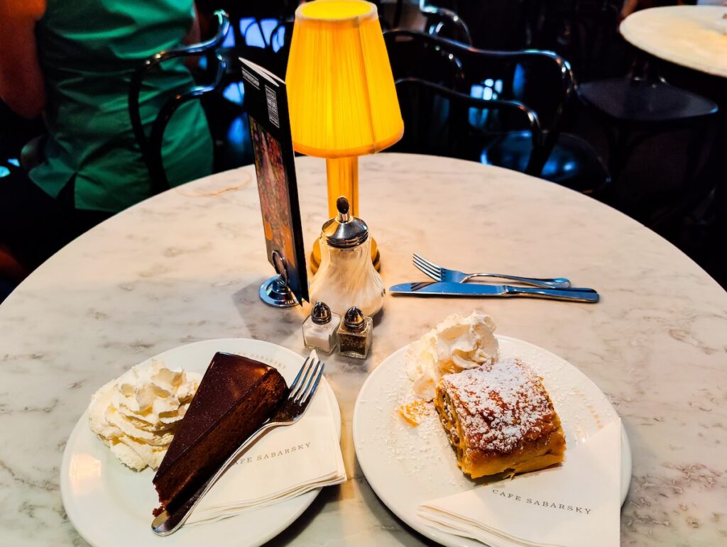 View of a white granite table with a small lamp with a lampshade and a plate with apple strudel and a plate with sachertorte. 