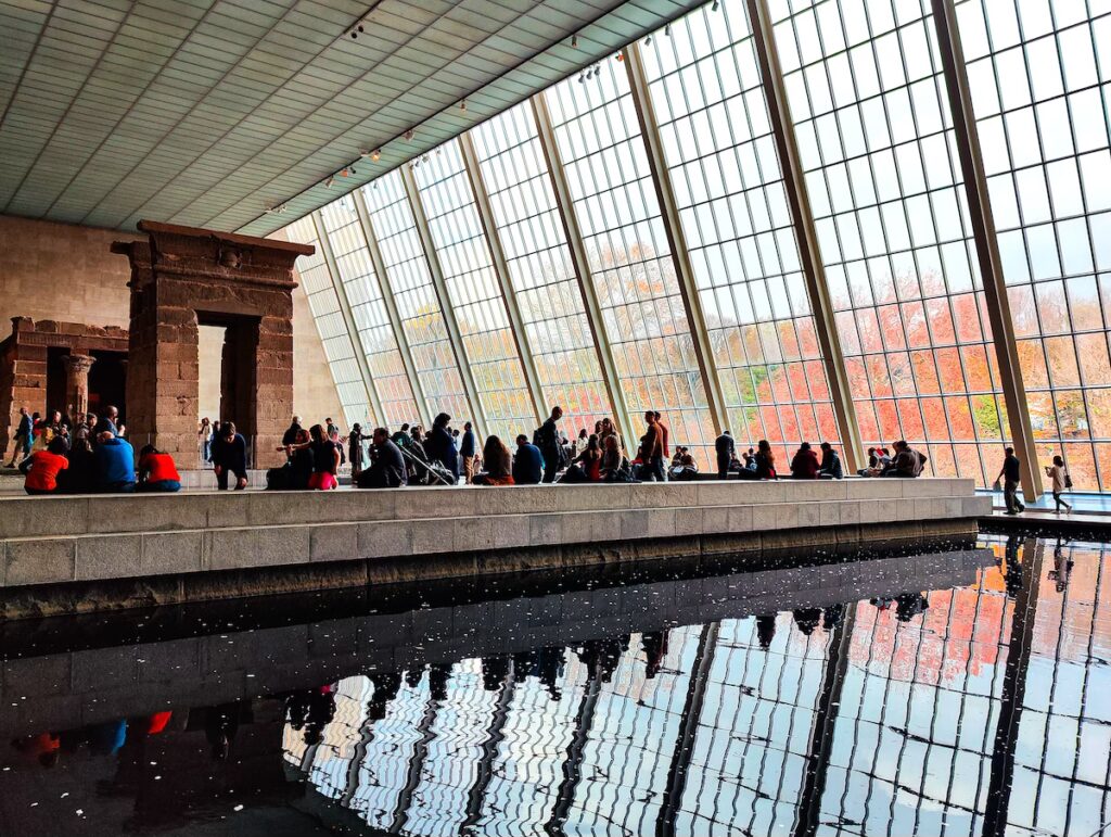 View of the Temple of Dendur at the Metropolitan Museum of Art. You can see a small temple in the back that is surrounded by a black pool and a wall of windows on the right. 