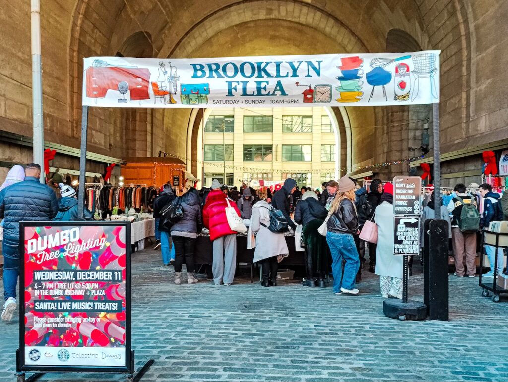 A view of people shopping beneath the Manhattan Bridge in Dumbo. You can see them standing beneath a huge sign that says, "Brooklyn Flea". 