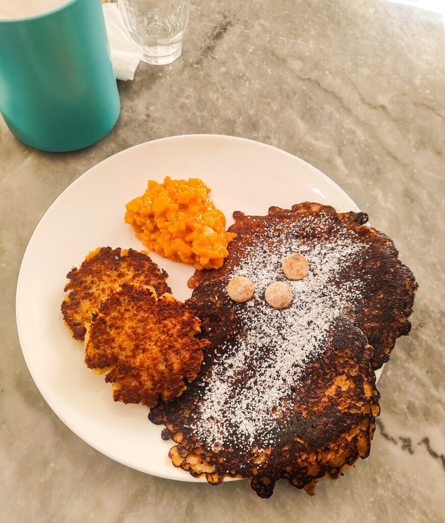 View of two soft scrambled eggs, brown griddle cakes with cinnamon butter and powdered sugar, and garbanzo bean sausages on a white plate with a turquoise cup on theft. 