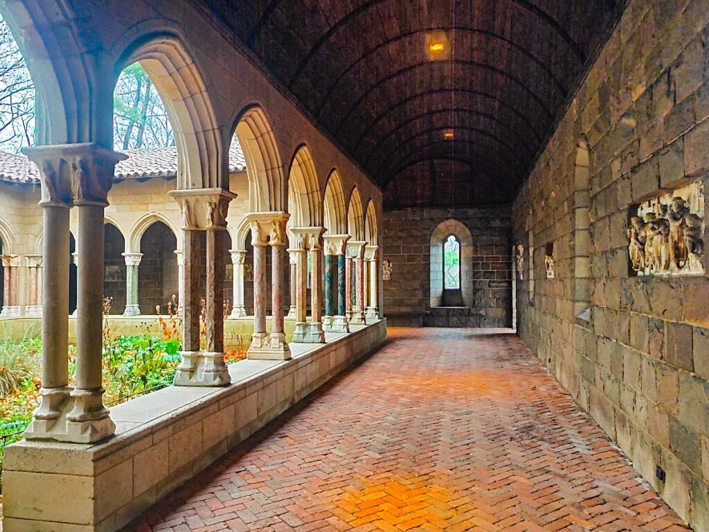 A beautiful view of the empty cloisters that surround a garden in the Cloisters MET location on a rainy day. You can see all the stonework in the building as you enjoy one of the best things to do in spring in NYC. 